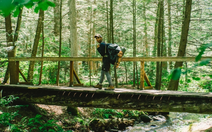 a young person carrying a backpack crosses a wooden bridge over a creek in a wooded area